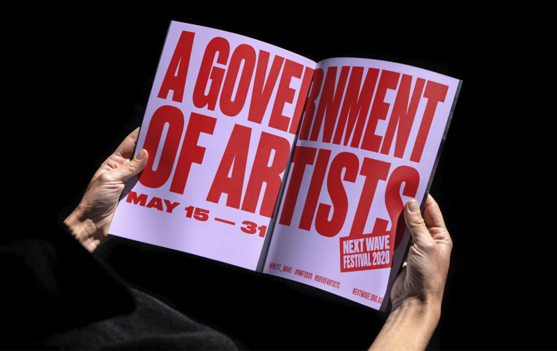 A person reading the Next Wave Festival, 2020 brochure. Red bold text on a pink page reading 'A Government of Artists'. May 15-31. Next Wave Festival 2020